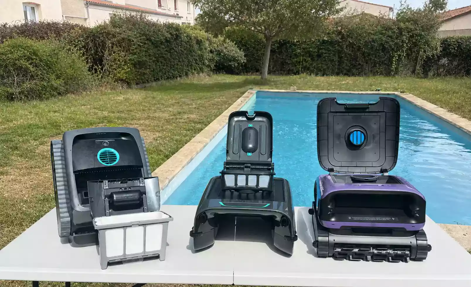 Trois robots piscine sans fil alignés sur une table au bord du bassin, paniers filtrants et trappes ouverts après test