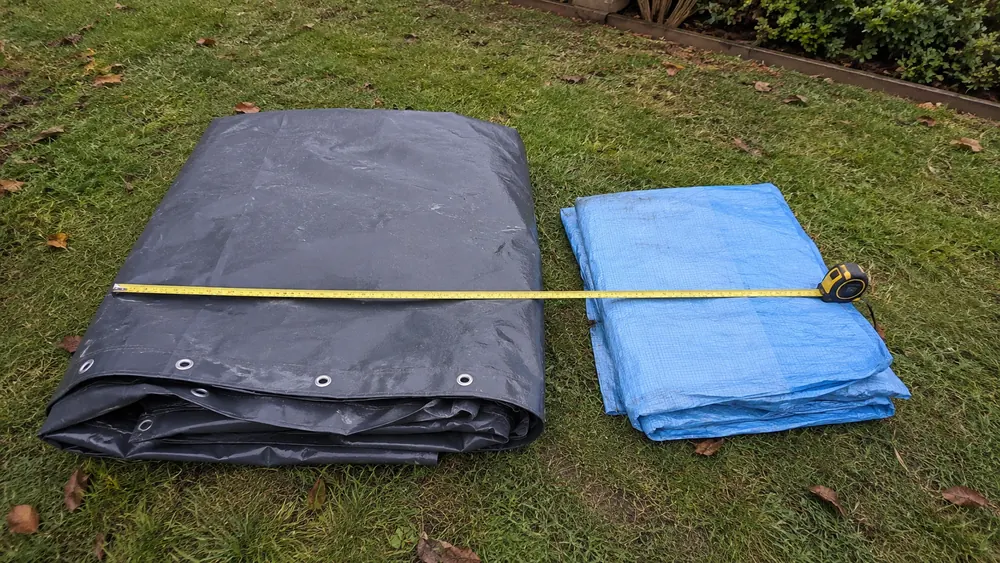 Deux bâches pliées côte à côte sur une pelouse, PVC armé gris et polyéthylène bleu, mètre ruban posé dessus