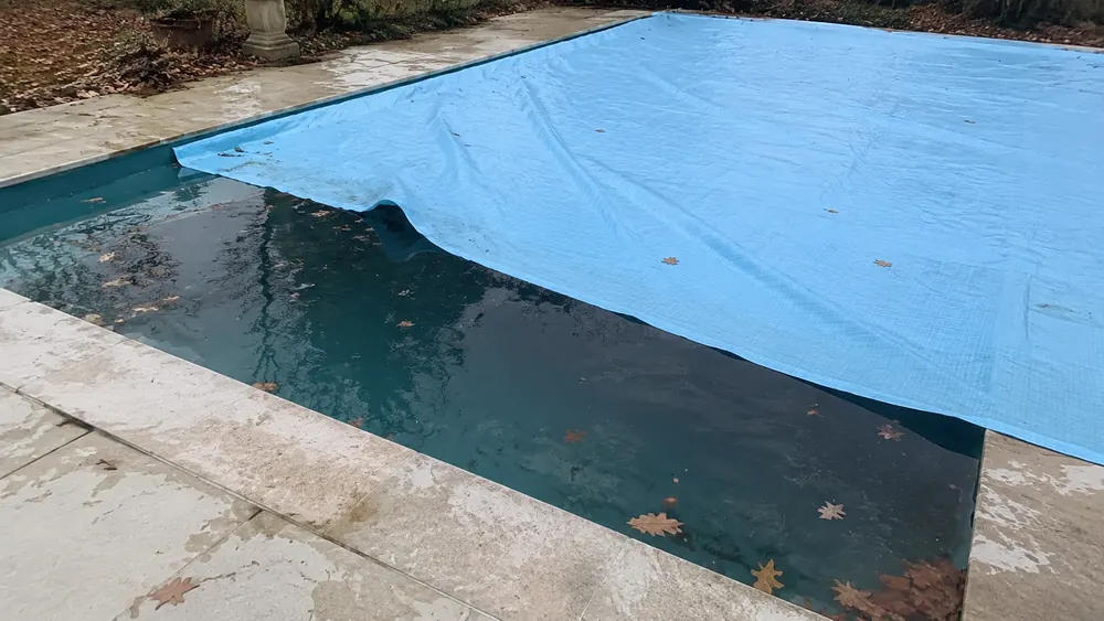 Bâche en polyéthylène bleu trop petite sur une piscine, angle du bassin découvert avec feuilles mortes sur l'eau
