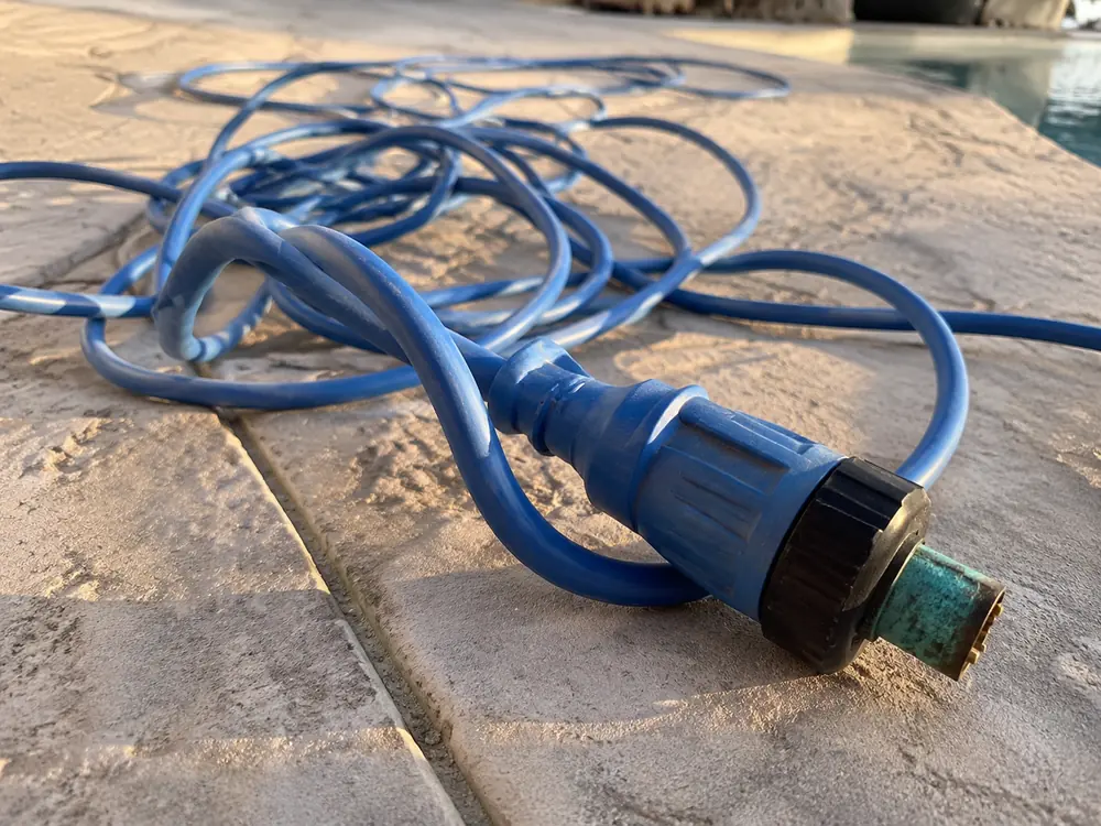 Câble bleu de robot de piscine enroulé sur une margelle, dont le connecteur métallique est couvert d'oxydation verte