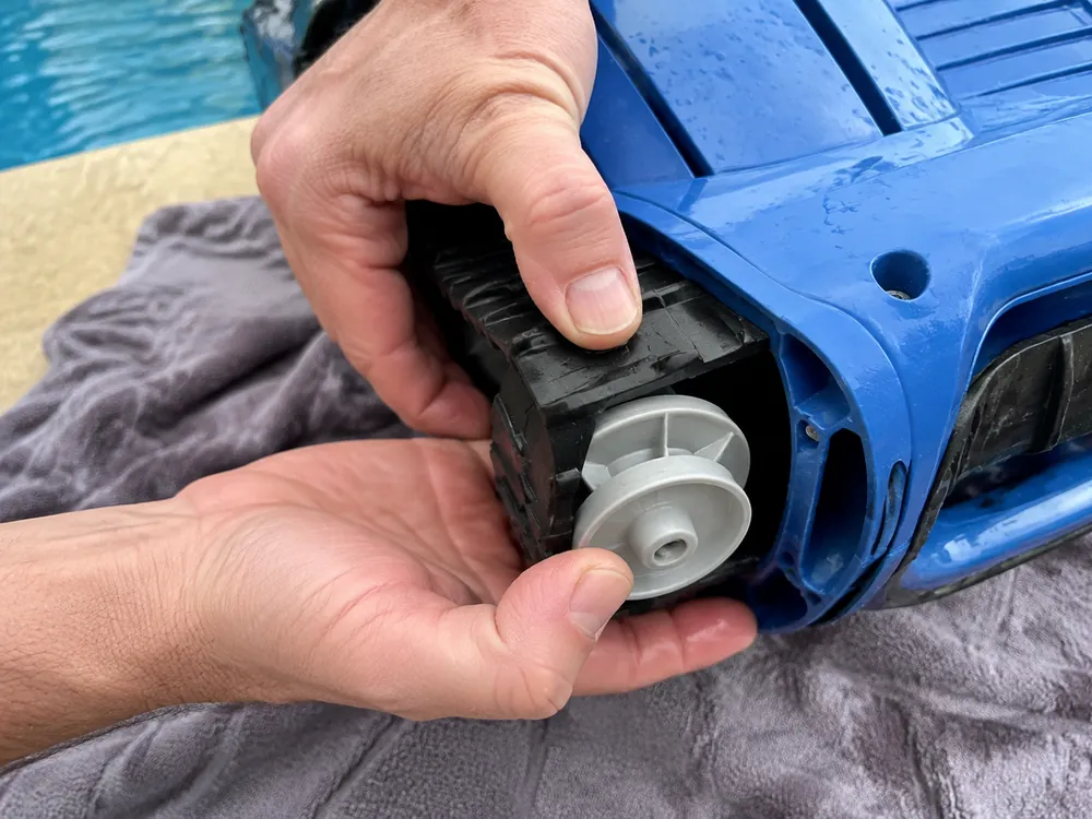 Deux mains en train de déclipser la chenille noire du galet gris d'un robot de piscine bleu posé sur une serviette au bord du bassin