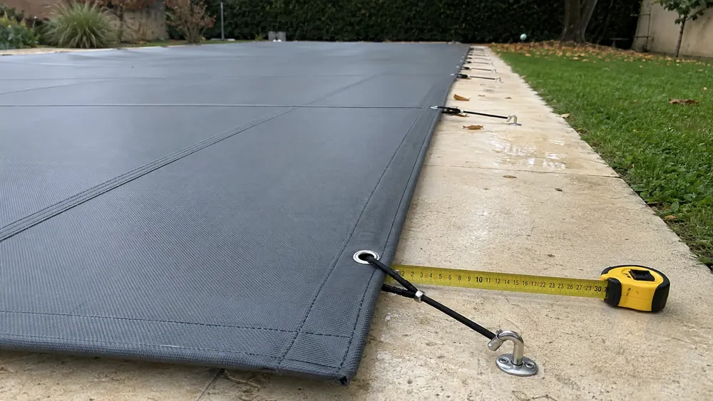 Mètre ruban mesurant le débord d'une bâche d'hivernage entre l'œillet, le sandow et le piton d'ancrage sur la margelle