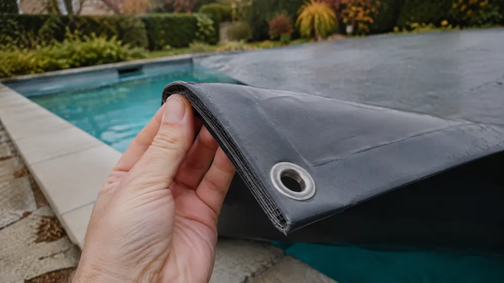 Coin d'une bâche d'hivernage tenu à la main, trame armée visible dans l'épaisseur et œillet inox
