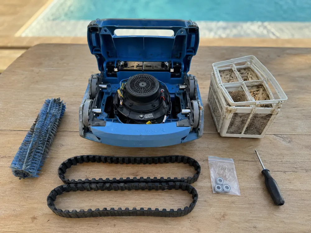 Robot de piscine bleu ouvert sur une table en bois, entouré de sa brosse, de ses deux chenilles, de son panier filtrant encrassé, d'un sachet de roulements et d'un tournevis